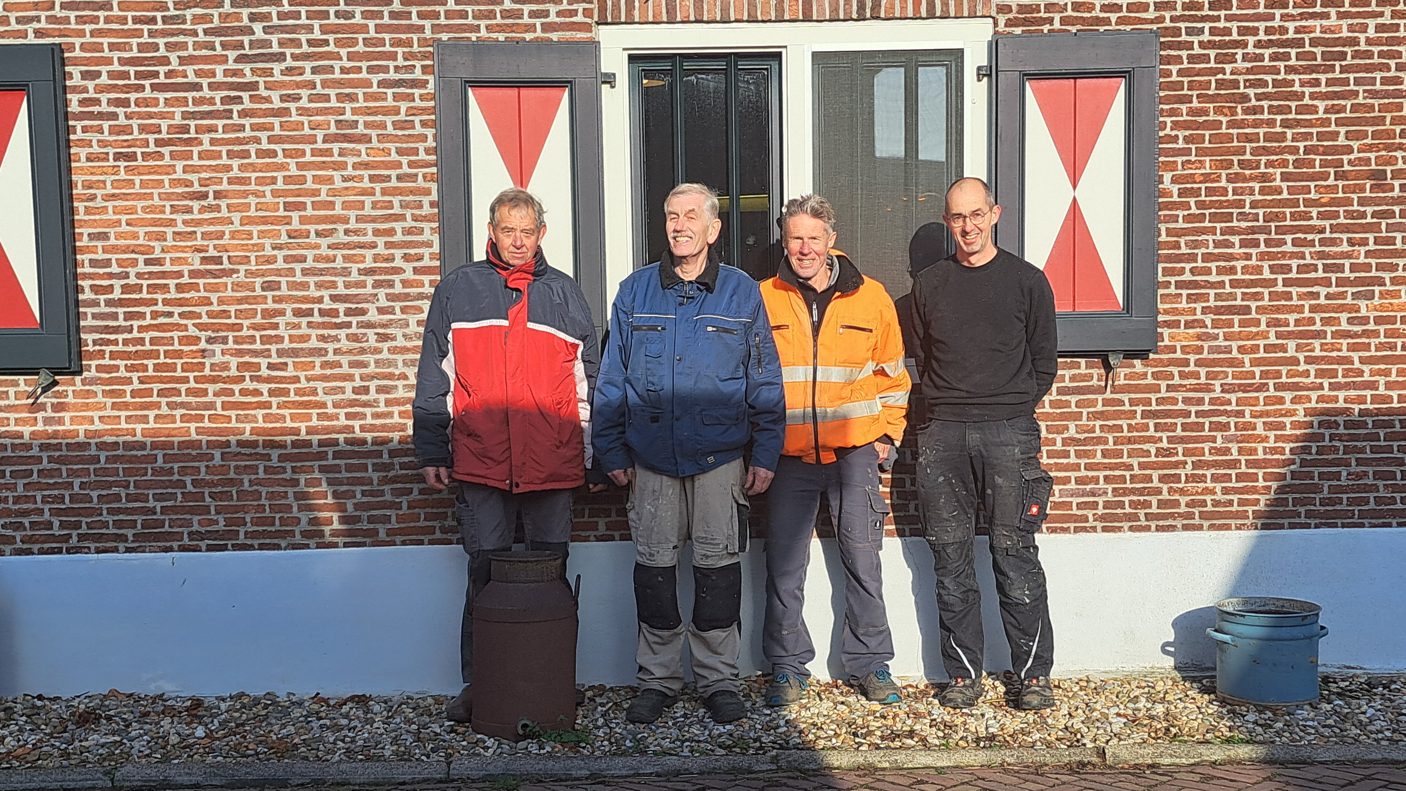 Twee mannen in blauwe overalls werken samen aan een kruiwagen in een buitenomgeving.
