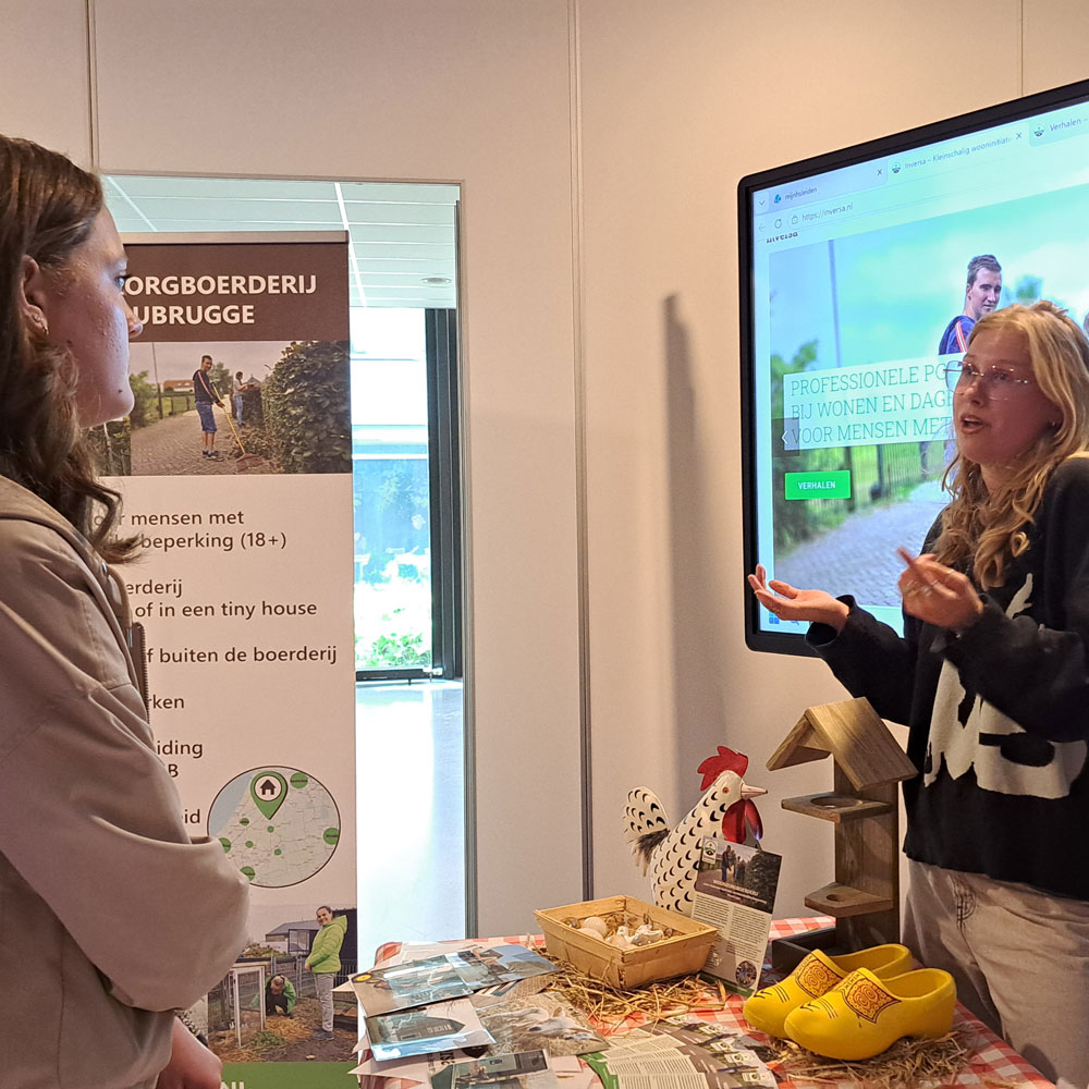 Twee vrouwen in gesprek aan een tafel met een groot scherm op de achtergrond.