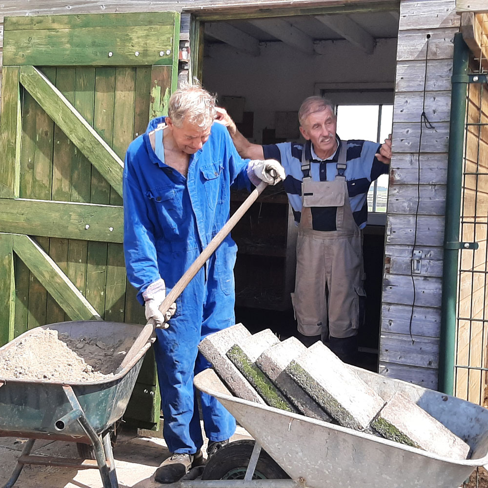Twee mannen in blauwe overalls werken samen aan een kruiwagen in een buitenomgeving.