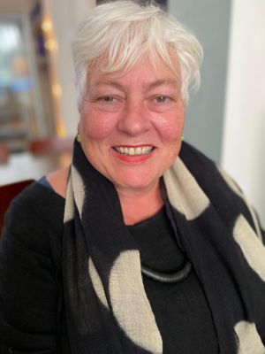 Een vrouw met wit haar draagt een zwart-wit sjaal en kijkt naar de camera met een vriendelijke glimlach.
